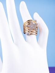 A close-up of a gold spiral-shaped ring with a large star adorned with rhinestones, worn on a white mannequin against a blue background