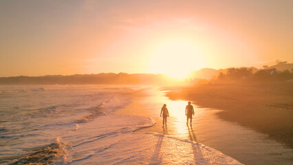 AERIAL: Beautiful view of a golden shining ocean beach with surfers strolling by