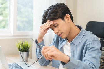 Exhausted, asian young businessman work at office desk, male took off eye glasses, headache from migraine, suffer on work hard, using laptop computer at office home, overtime job. Health care concept.