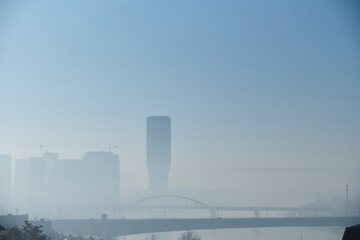 Atmospheric urban landscape showing city skyline partially obscured by dense fog, with bridge crossing river in foreground