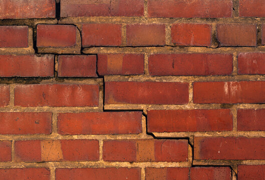 Cracked brick wall showing structural damage. Step crack pattern in exterior brick wall. Foundation crack in red brick house wall. Close up of masonry crack in brick facade.