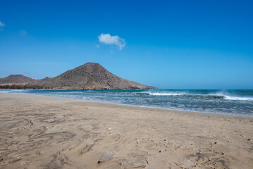 A view of Playa de los Genoveses, Almeria, Spain, with its golden sand, turquoise waters, and a hill in the background under a clear blue sky. A peaceful, unspoiled natural landscape