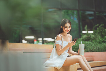 Happy young Asian woman sitting on steps using a smartphone and holding a reusable tumbler with a white tote bag in a modern urban environment