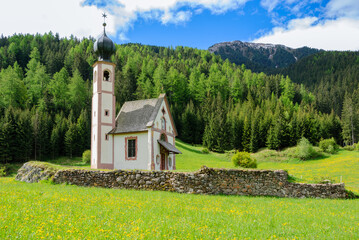 Small alpine chapel in a green meadow surrounded by forested mountains under a blue sky, peaceful rural landscape in spring.