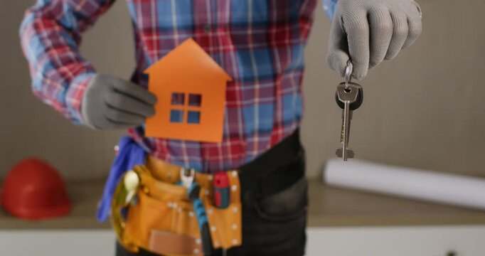 A worker lowers a bunch of keys to a new apartment and house into the frame, and in the background he holds a stencil of a house in his hand.