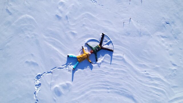 Top view aerial shot of a man and woman lying on fresh snow and making snow angel shapes. Winter outdoor activity, happiness, freedom and seasonal lifestyle concept with copy space.