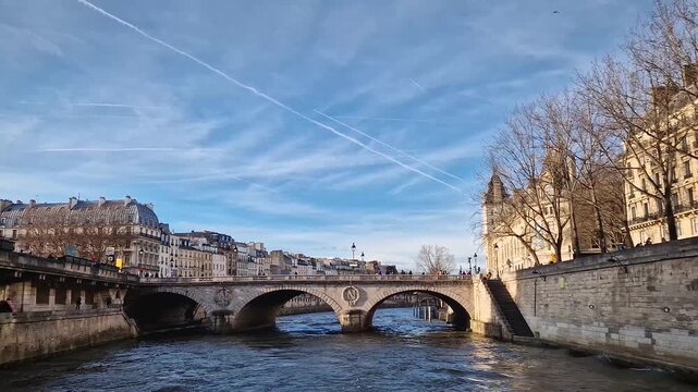 View from a cruising boat along the Seine River to the historic Pont au Change bridge. The stone arches feature the imperial N insignia. Bare trees and classic Parisian buildings line the riverbanks