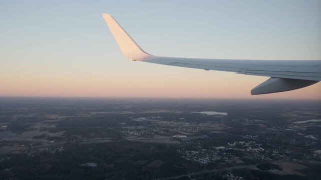 Airplane wing with aerodynamic winglet flies over rural fields and forests during pastel sunrise. Commercial flight journey view showing calm morning sky and scenic ground landscape below.