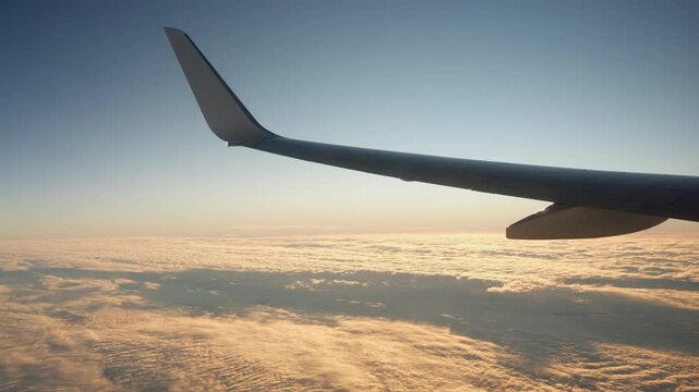 Commercial airplane wing with winglet flies above thick layer of golden clouds. High altitude flight journey view showing orange sunrise sunlight on majestic cloudscape.