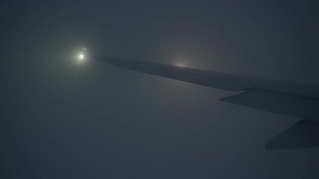 Commercial airplane wing flies through dense dark fog at night with flashing strobe and red navigation lights. Aircraft wingtip visible in heavy mist and low visibility weather conditions.