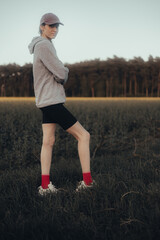 Vertical portrait of a young woman in a gray sweatshirt and hat, standing confidently in a field at dusk with a forest in the background and a place for text