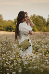Young woman in white lace dress with green handbag posing in blooming daisy field during golden hour, cottagecore vertical photo with blurry background of nature and free space for text