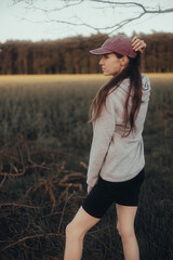 A young active woman in a gray hoodie and a burgundy cap standing on a meadow at sunset looking towards the forest in a vertical frame
