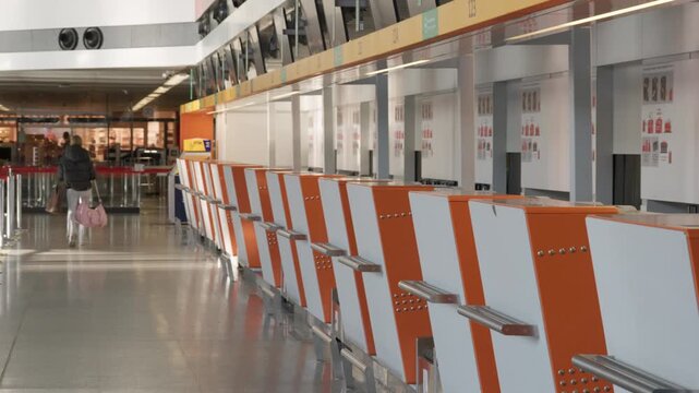 Row of empty check-in desks at Warsaw Chopin Airport terminal. Unmanned passenger baggage drop-off counters with orange branding standing in departure hall while traveler walks in background.