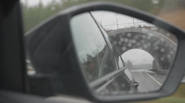 Reflection of receding wet asphalt road in car side wing mirror. View of highway traffic and bridge arch through window with raindrops, symbolizing life journey, hindsight and moving away.