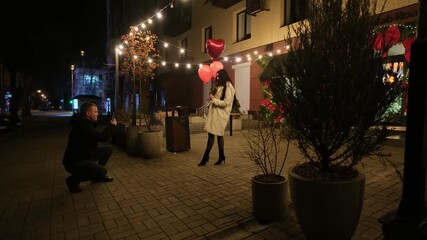 Young romantic couple enjoying a city date at night, with a man taking a smartphone picture of his beautiful girlfriend holding red heart-shaped balloons under festive string lights - Powered by Adobe