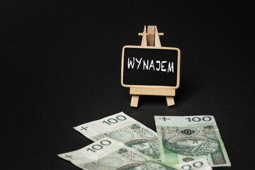 Black wooden writing board on a black background with the inscription "Wynajem", in the background scattered Polish złoty PLN banknotes (selective focus)