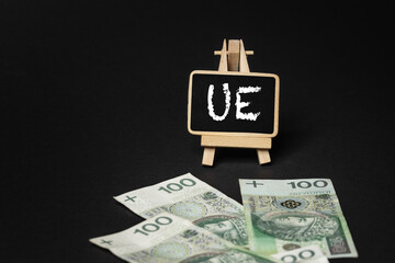 Black wooden writing board on a black background with the inscription "UE", in the background scattered Polish złoty PLN banknotes (selective focus)
