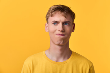 Fototapeta premium Young man in a yellow shirt against a yellow backdrop shows confusion and curiosity, a facial expression of doubt and hesitation after unexpected news or a tough decision.