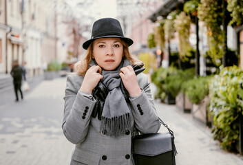 Business woman in grey suit, scarf, gloves and black hat standing on city street, adjusting scarf . Urban lifestyle, cold season, stress, career and emotional concept.