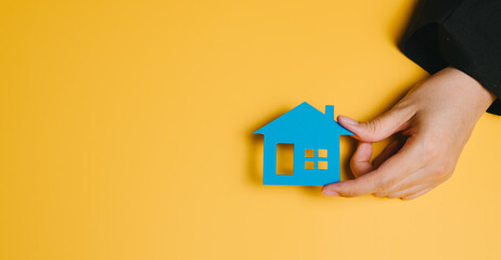 Buying a house concept. Blue house model held by hand on yellow background symbolizing real estate...