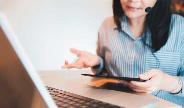 Asian businesswoman wearing a headset engaging in an online meeting, using a digital tablet and laptop. Ideal for concepts like remote work, virtual collaboration, and professional communication. - Powered by Adobe