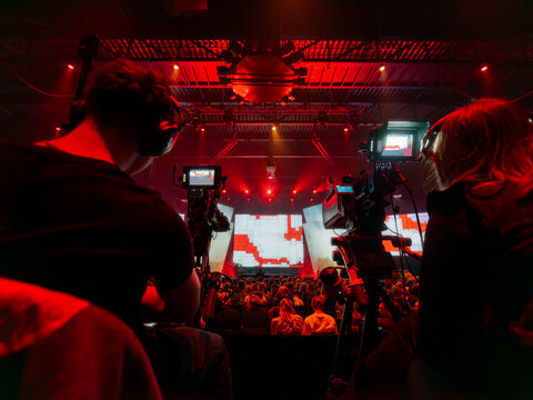 Multiple broadcast cameras filming large venue event with red stage lighting and screens