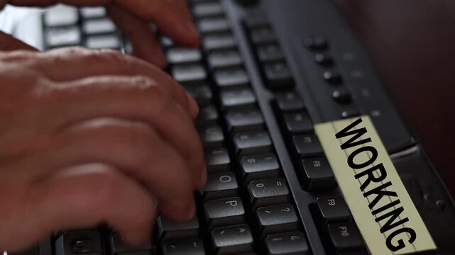 Un primer plano de un teclado de una computadora con las manos de una mujer adulta y una nota amarilla con la palabra trabajando o trabajo