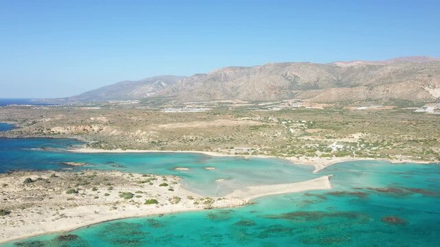 Stunning aerial view of Elafonisi beach with clear turquoise waters, sandy shores, and rugged mountains in Crete, Greece. The landscape features a unique sandbar and natural coastal beauty.