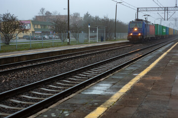 Freight train with containers passing through a railway station on a foggy morning. Industrial rail transport scene with empty platform, tracks and urban infrastructure