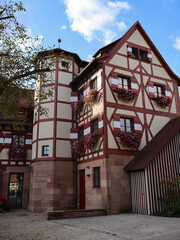 Traditional timber-framed building in Nuremberg with red trim and colorful flower boxes, set beside a cobblestone plaza. Bright sky adds warmth to this picturesque European village scene.