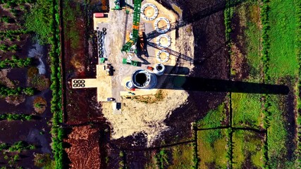 An overhead view of a wind turbine construction site shows a tall concrete tower section, machinery, circular foundation parts, and access paths surrounded by patterned peat fields and green patches.