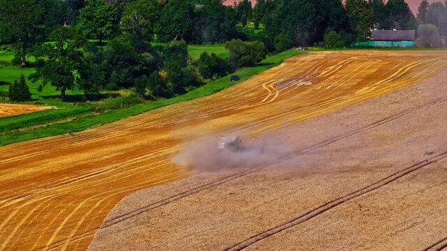 A wide farm slope shows golden crop rows with a machine raising dust as it moves, while green trees form a contrasting band at the top of the scene.