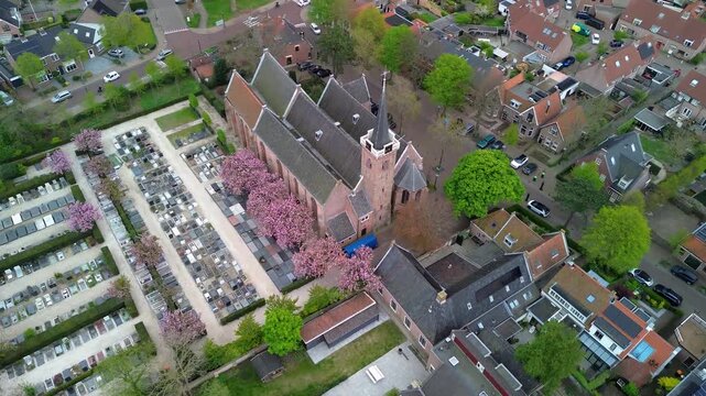Aerial view of church and cemetery steeple towers over rows of gravestones and blooming pink trees, surrounded by red roofed homes and quiet streets that form a serene, harmonious townscape.