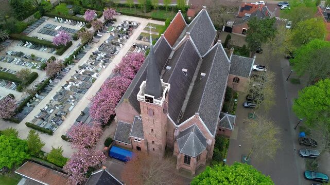 Aerial view of church and cemetery steeple towers over rows of gravestones and blooming pink trees, surrounded by red roofed homes and quiet streets that form a serene, harmonious townscape.