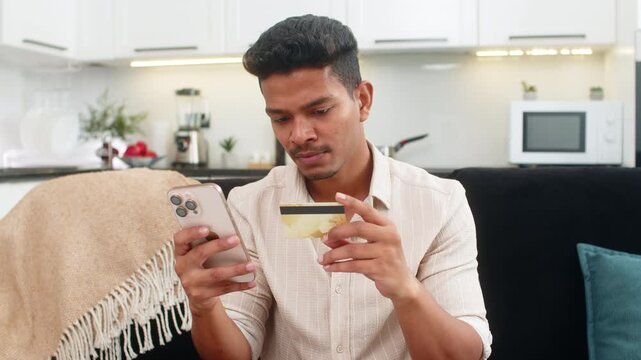 Indian man at home orders delivery on smartphone enters card data checks address and confirms payment. Arabian guy on sofa shows thumbs up approval order confirmed waits courier tracking notification