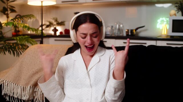 Young woman at night home wears headphones hears song starts seated dance laughs cheers yeah. Caucasian girl on sofa at evening makes rock hand signs nods to beat smiles shows joy fun energy music oh.