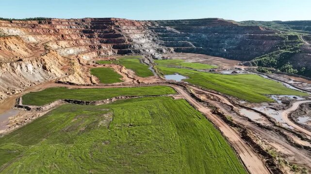 Expansive mine site showing terraced rehabilitation with green growth and muddy tracks, Aerial view of a vast open-pit mine undergoing reforestation efforts with vibrant green vegetation.