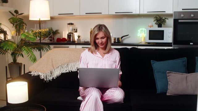 Adult woman at night home opens laptop starts project types fast checks tasks progress smiles with focus. Caucasian girl on sofa at evening marks first tasks complete updates board notes work tracker.