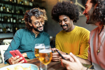 Three diverse young adult friends using mobile phone sitting on table at bar. Technology and male friendship concept.