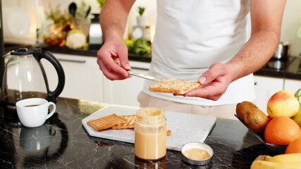 Preparing breakfast spreading peanut butter on toast in home kitchen for quick morning meal. Close up hands using knife from jar making snack with coffee for energy and healthy nutrition before work.