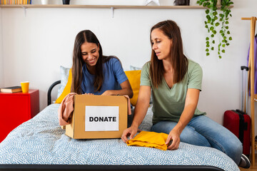 Caucasian teenage girl and her mother sitting on the bed, organizing clothes in a cardboard box to donate. Concept of generosity, family values, and social responsibility at home.