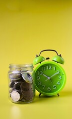 Green alarm clock and jar filled with coins on yellow background