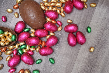 Pile of colourful foil wrapped chocolate easter eggs in pink, red, gold and green. Large halves of a brown milk chocolate egg have mini eggs inside, on a grey wooden background.