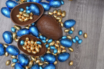 Pile of colourful foil wrapped chocolate easter eggs in blue, and gold. Large halves of a brown milk chocolate egg have mini eggs inside, on a grey wooden background.
