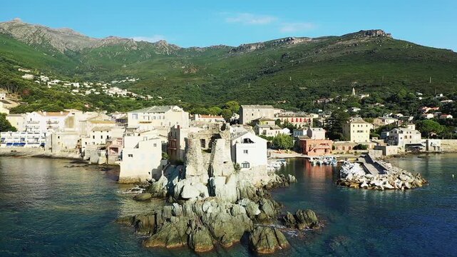 Aerial view of the picturesque seaside village of Erbalunga, Corsica, with historic stone buildings, a small harbor, and lush green mountains in the background.