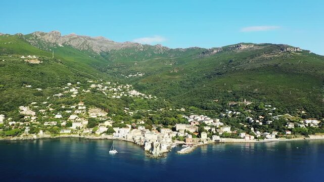 Scenic aerial view of Erbalunga village nestled on the Mediterranean coast with lush green hills and clear blue water, under a bright sky.