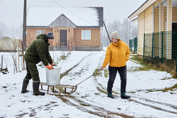 Manual waste removal in rural area on sled to garbage container in winter time, mature couple works together.