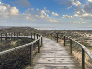 A wooden boardwalk curves across sandy coastal dunes covered with low vegetation, offering a panoramic view of the Furadouro beach and Atlantic Ocean under a bright, cloudy sky in Ovar Portugal