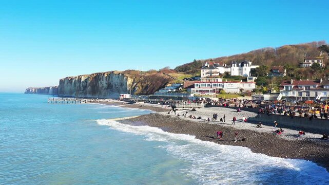 Scenic view of the pebble beach and dramatic white cliffs at Veules les Roses, France, with people enjoying a sunny day by the sea. Picturesque coastal village with traditional houses and clear blue